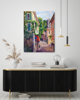 A canvas painting of a sunlit narrow street flanked by colorful buildings with balconies and greenery, displayed above a modern black and gold console table in a minimalist room.