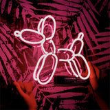 Pink neon light shaped like a balloon dog being held by two hands against a patterned dark red and black wall.
