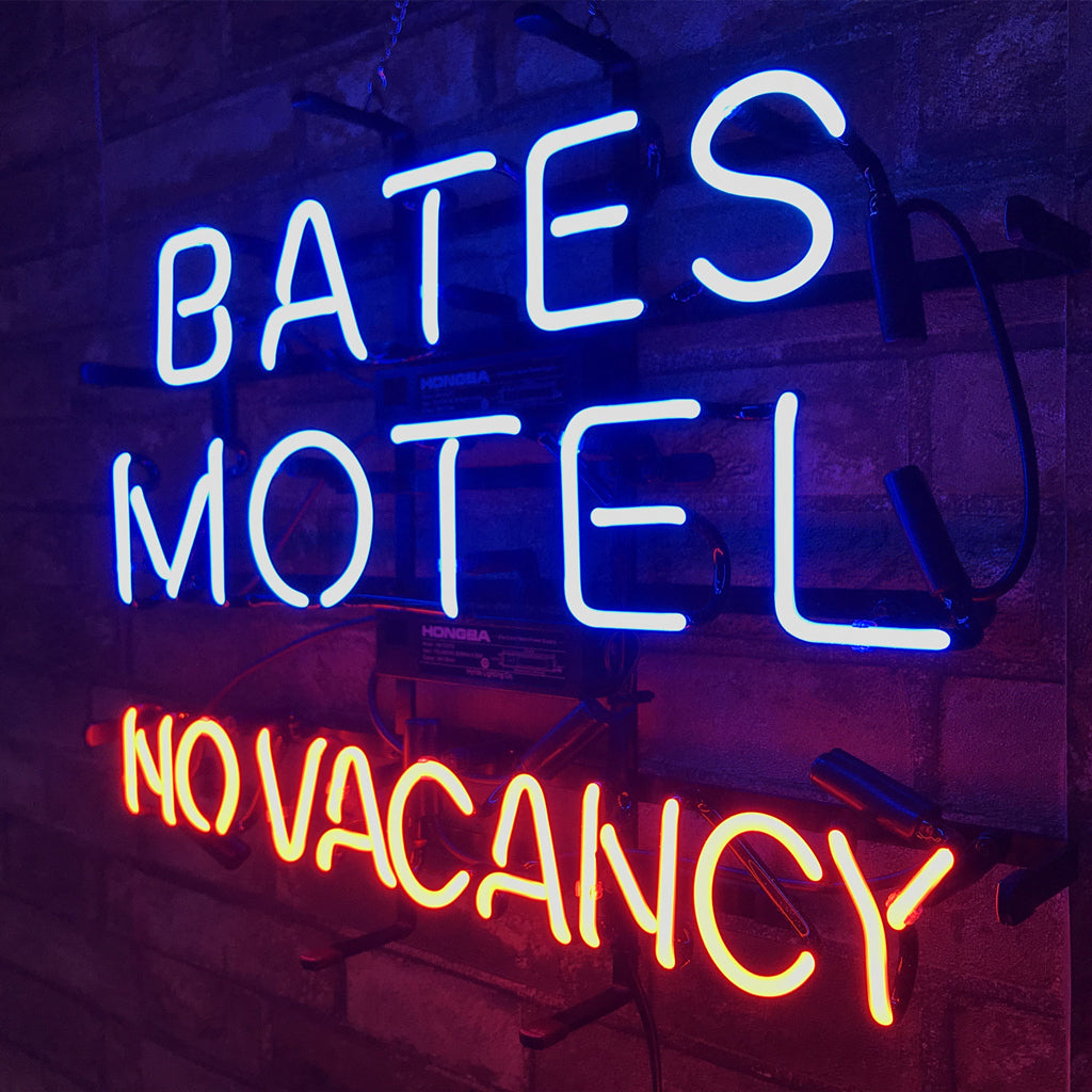 Neon sign with bright blue letters spelling 'BATES MOTEL' above red letters reading 'NO VACANCY' mounted on a dark brick wall.