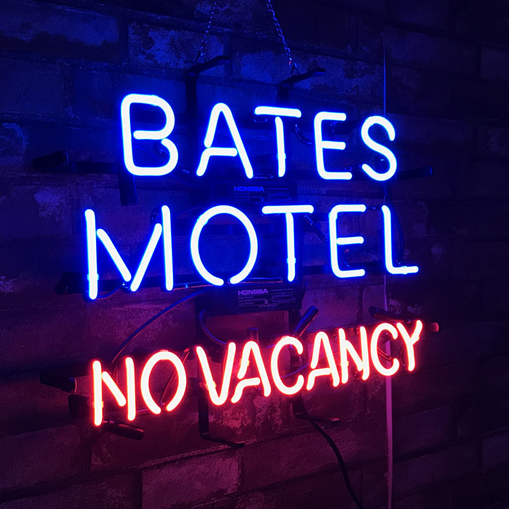 Neon sign with blue text 'BATES MOTEL' above red text 'NO VACANCY' mounted on a dark brick wall and lit in dim lighting.