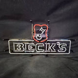 Neon light sign with white glowing text reading 'BECK'S' inside a rectangular outline and a red shield emblem above it against a dark background.