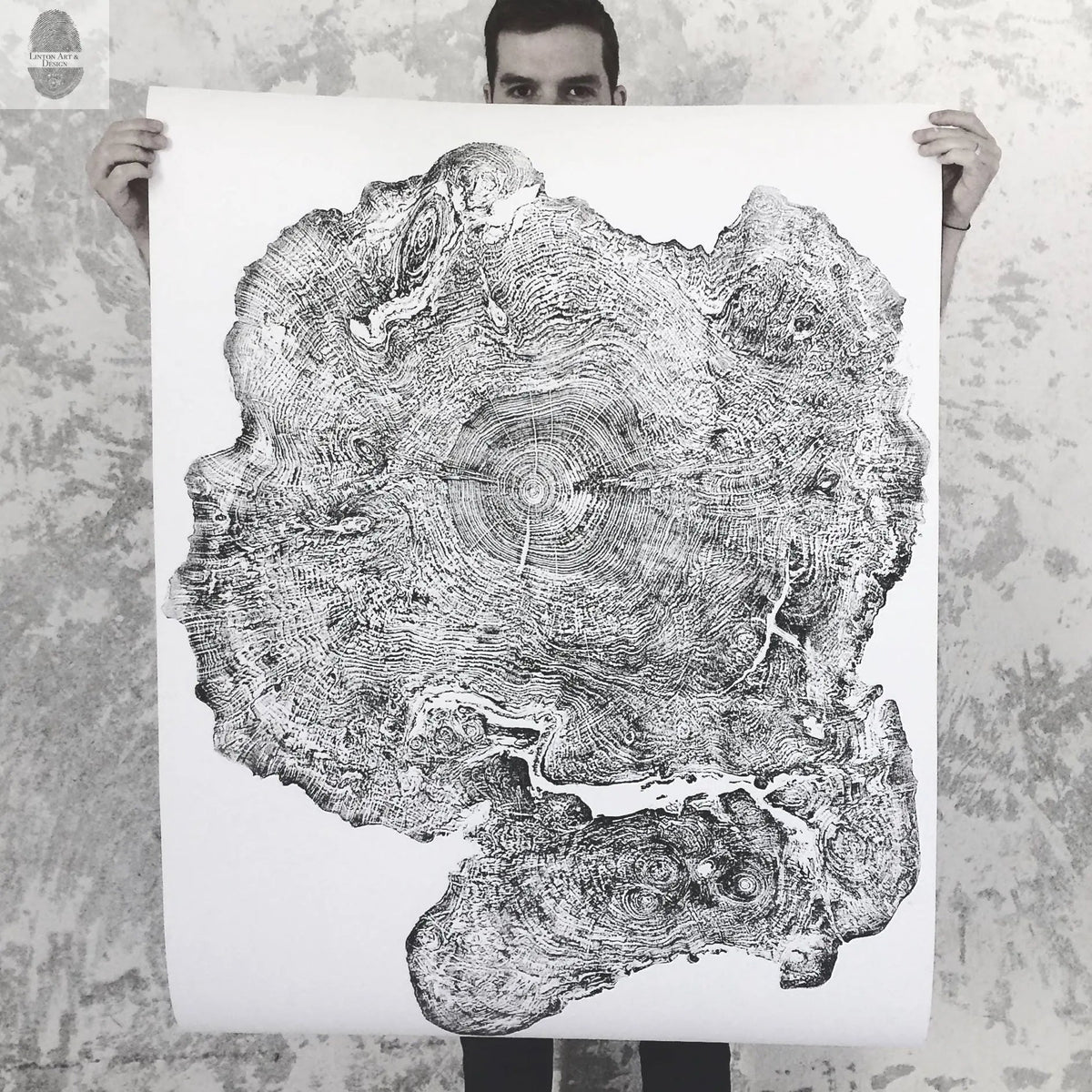 Black and white hand-pressed print of a Big Leaf Maple tree ring showing detailed concentric growth rings and natural bark edge held by a person against a mottled wall.