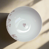 White ceramic bowl with delicate brown floral branches on the rim and a small heart with initials inside the center, shown from above with soft shadow lines.