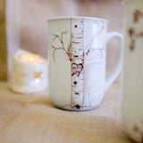 White ceramic mug featuring a delicate birch tree illustration with a carved heart containing initials K+T and the date 1-23 on the tree trunk.