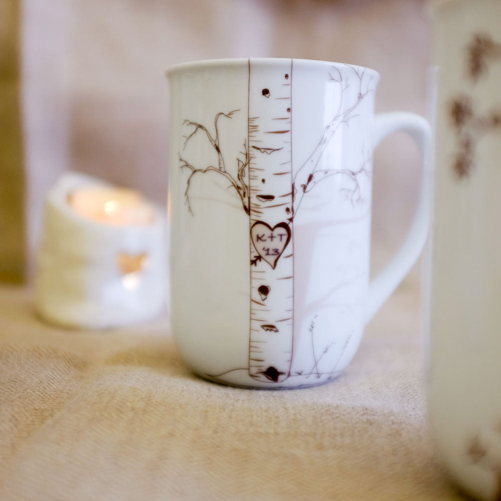 White ceramic mug featuring a delicate birch tree illustration with a carved heart containing initials K+T and the date 1-23 on the tree trunk.