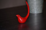 Small glossy red bird-shaped ring holder with a tall curved tail, placed on a dark wooden surface near a gray textured pot in the background.