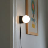 Wall-mounted sconce with a matte black square base and a glowing white spherical bulb, next to a colorful abstract artwork in a light wooden frame.