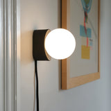 Wall-mounted black metal sconce with a luminous white glass globe, shown next to a framed colorful abstract print on a white wall.