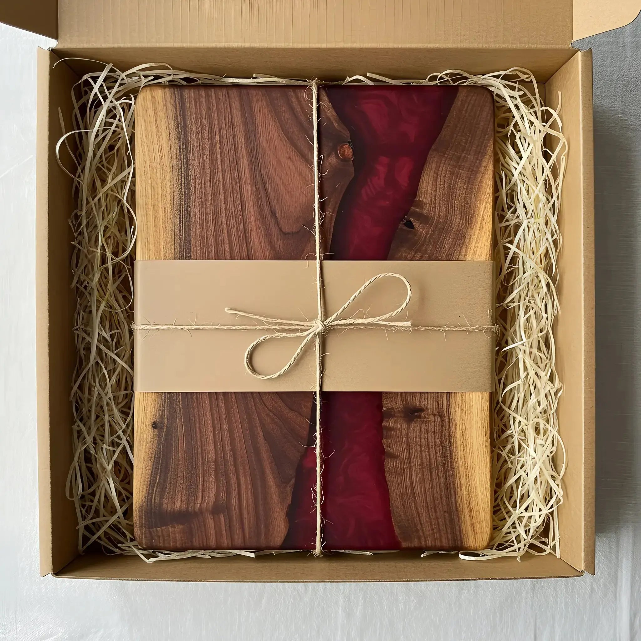 a charcuterie board carefully packaged for shipping. The board is made from live edge black walnut with a smooth, polished finish and a deep merlot red epoxy river running through the center. It’s wrapped in kraft paper and tied with natural twine for a handmade, artisan feel. Matte black metal handles are attached at each end