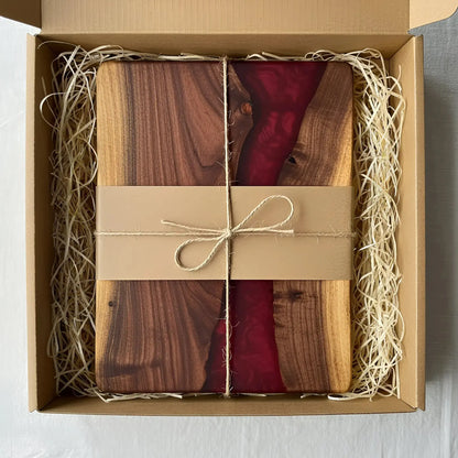 a charcuterie board carefully packaged for shipping. The board is made from live edge black walnut with a smooth, polished finish and a deep merlot red epoxy river running through the center. It’s wrapped in kraft paper and tied with natural twine for a handmade, artisan feel. Matte black metal handles are attached at each end