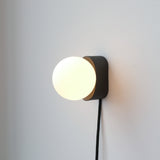Wall-mounted black rectangular base with a glowing white spherical light globe and a black fabric-covered cord hanging below, set against a plain white wall.