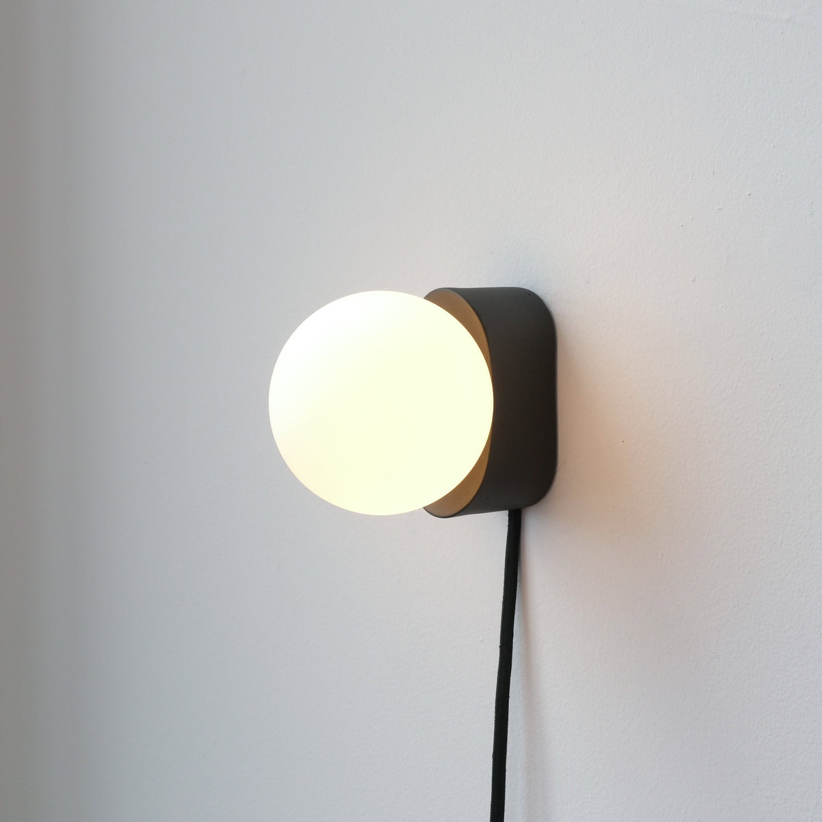 Wall-mounted black rectangular base with a glowing white spherical light globe and a black fabric-covered cord hanging below, set against a plain white wall.