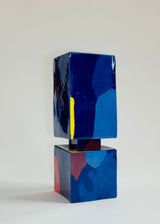 Glossy ceramic sculpture with a smaller blue cube atop a larger cube featuring red and blue abstract patches, separated by a thin dark rectangular base.