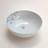 White ceramic bowl with a blue heart design at the center and delicate blue floral patterns along the inner rim on one side.