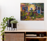 Impressionist painting of a yellow house with blue windows and a wooden gate, surrounded by lush green foliage and pink flowers, displayed on a white wall above a wooden cabinet.