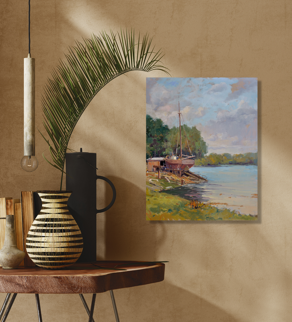 Canvas print depicting a boat on a launch ramp beside a river under a cloudy sky, hanging on a beige wall above a wooden table with vases and a palm leaf.