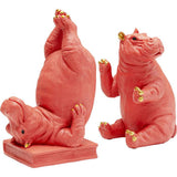 Two pink hippo figurines with gold-tipped ears, paws, and toes, one sitting upright and the other lying on its back with legs raised on a book base.