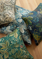 Four decorative pillows with intricate floral patterns in muted green, blue, and beige tones on a light wood floor, featuring leafy and flower motifs.
