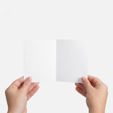 Two hands holding a small open blank white card against a plain white background, showing the inside spread with no text or graphics.