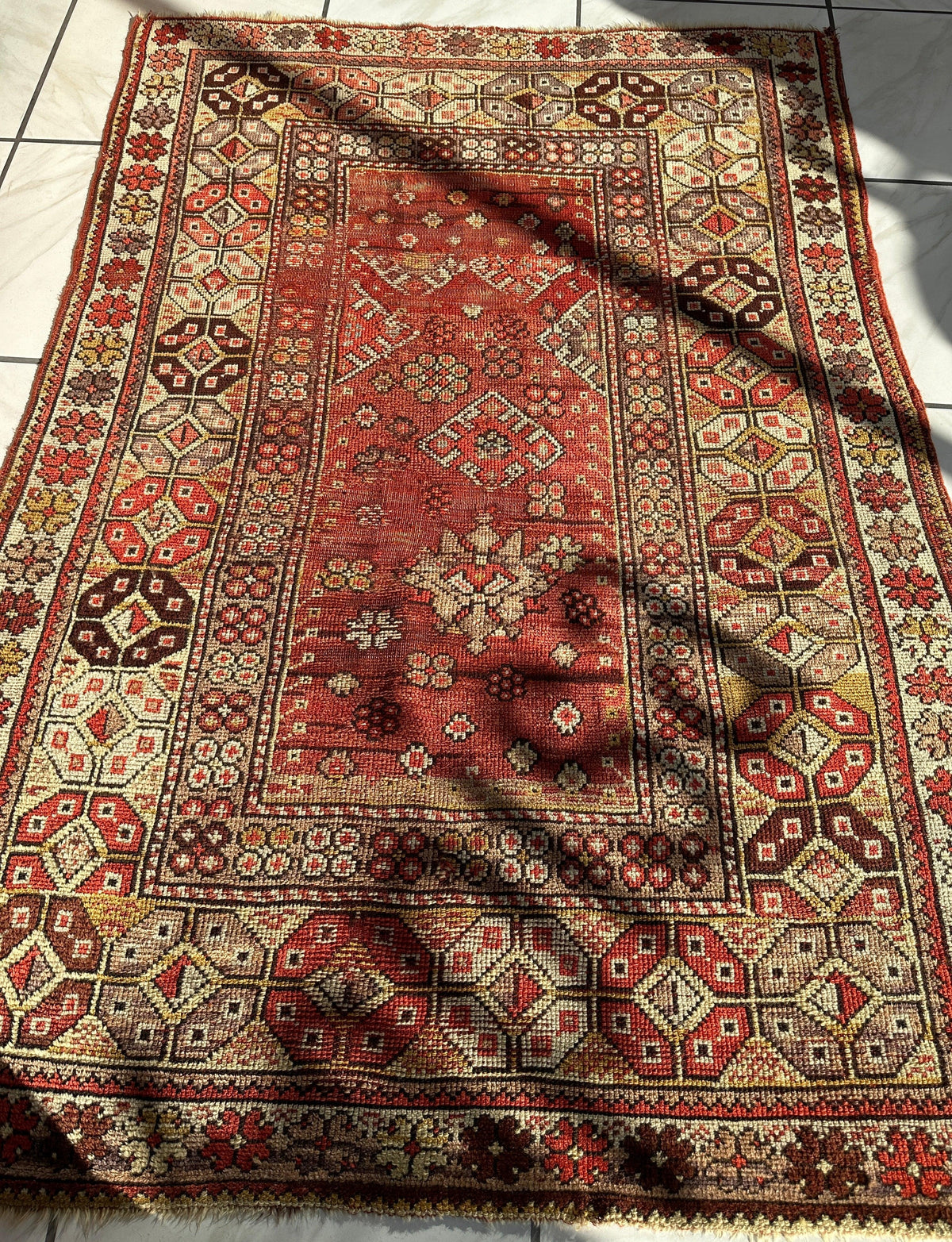 Rectangular antique Turkish prayer rug with distressed red central field featuring geometric floral motifs, surrounded by intricate beige and brown patterned borders and a repeating hexagonal design.