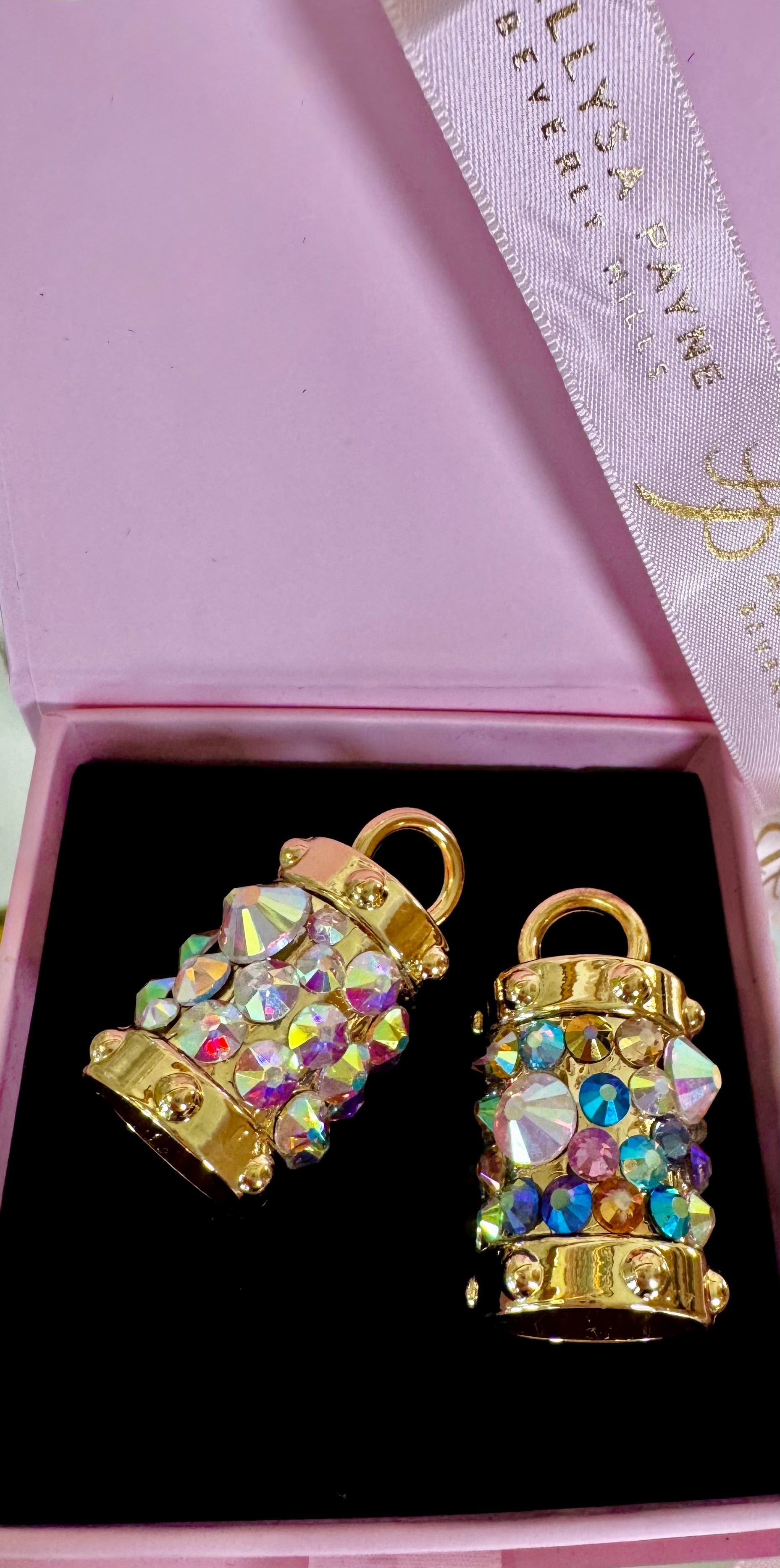 Two gold-tone bell charms adorned with iridescent Swarovski crystals, displayed on a dark surface.