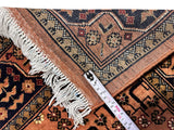 Corner of a hand-knotted Afghan Bukharayi rug in peach and navy with geometric and floral patterns, showing white fringed edges and a measuring tape for scale.