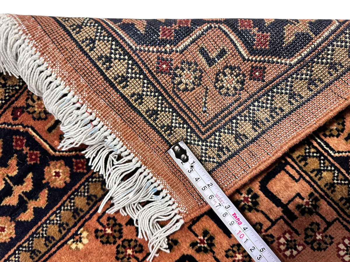 Corner of a hand-knotted Afghan Bukharayi rug in peach and navy with geometric and floral patterns, showing white fringed edges and a measuring tape for scale.