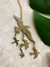 Brass bird ornament with one large bird shape and four smaller birds hanging below, displayed on a beige marble surface with green leaves nearby.