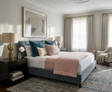 Bedroom with blue upholstered bed featuring coral pink quilt and patterned pillows, flanked by dark wood nightstands with gold lamps, soft natural light filling the room.