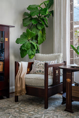 Wood-framed armchair with patterned beige and gray cushions and a decorative pillow, draped with a beige throw, set beside a fiddle leaf fig plant near a window.
