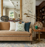 Tan velvet sofa with three decorative pillows in cream, green botanical print, and blue velvet, set against a floral bird wallpaper and large framed mirror reflecting a chandelier.
