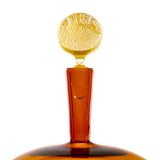 Tall amber glass bottle with a wide base and a round, textured golden glass stopper, shown upright against a white background.