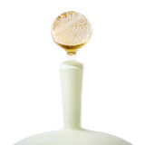 Curved white glass bottle with a textured amber spherical stopper, shown in close-up against a white background.