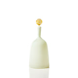 Opaque pale green wide bottle with a narrow neck topped by a textured amber round stopper, shown against a white background with soft reflection.