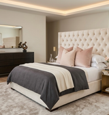 Bedroom with cream tufted headboard and bed layered in grey and cream bedding, accented by two blush pink spotted pillows, against beige walls and soft lighting.