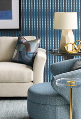 Abstract curved patterned indigo pillow on cream sofa next to blue textured chair, round glass and brass side table with beige lamp and gold decorative rings