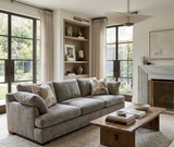 Neutral-toned living room with a gray sofa adorned with parchment and patterned pillows, a rustic wooden coffee table, and a built-in shelf with art pieces.