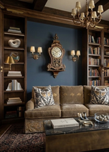 Classic sitting area with a beige velvet sofa adorned with two gold and grey patterned pillows, a dark bronze coffee table, and an ornate antique clock on a navy blue wall.