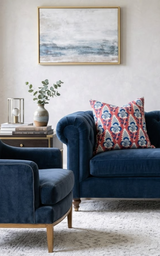 Red pillow with blue and white floral pattern on a dark blue velvet sofa, next to a matching armchair and small side table with books and a vase with greenery.