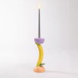 Candlestick shaped like a yellow banana attached to a peach-colored base with a purple holder supporting a tall lit gray candle