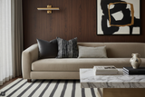 Modern beige sofa with four decorative pillows in black, white, and patterned designs against a dark wood paneled wall with abstract black and gold artwork.