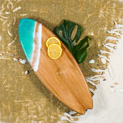 Wooden surfboard-shaped charcuterie board with blue resin accent, holding cheese and fruit on a patio table