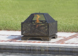 Square metal fire pit with geometric cutout patterns and mesh screen on a brick and stone patio next to a pool, flame visible inside.