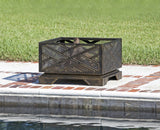 Square black metal fire pit with geometric cutout patterns on a raised base, placed on a brick-bordered concrete patio near a grassy lawn and pool edge.