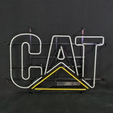 Neon sign spelling 'CAT' with white letters and a yellow triangular underline on a black metal frame against a dark background.