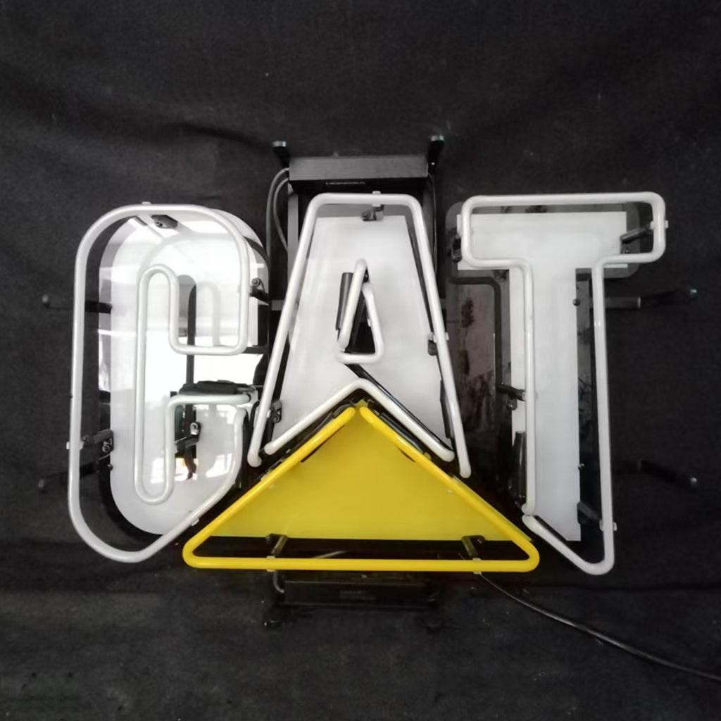 White neon letters spelling 'CAT' with a yellow triangular neon shape beneath the letter A, mounted on a black metal frame