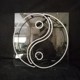 White neon light shaped as a Yin Yang symbol mounted on a square transparent backing, photographed against a dark background with industrial reflections.