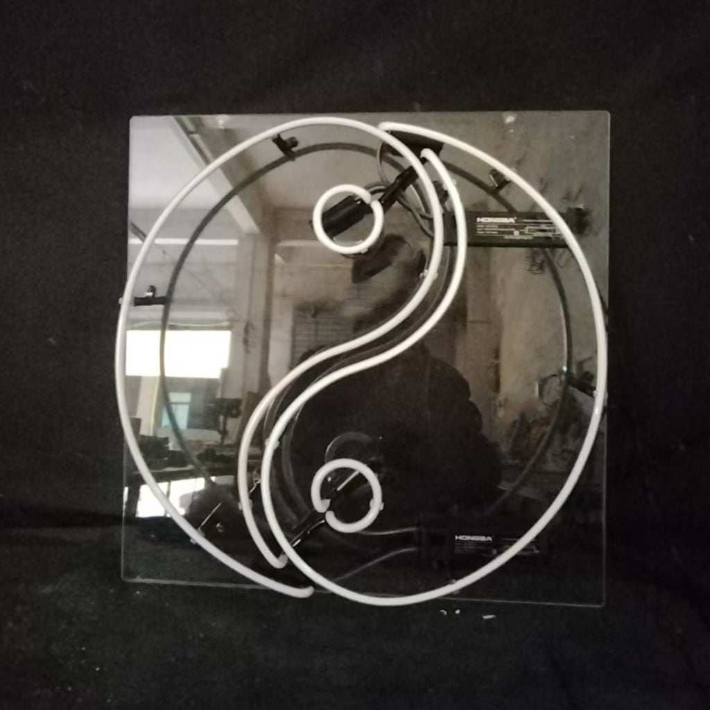 White neon light shaped as a Yin Yang symbol mounted on a square transparent backing, photographed against a dark background with industrial reflections.