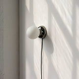 Wall-mounted sconce with a round black metal base and a frosted white glass globe, casting a soft shadow on a sunlit white wall.