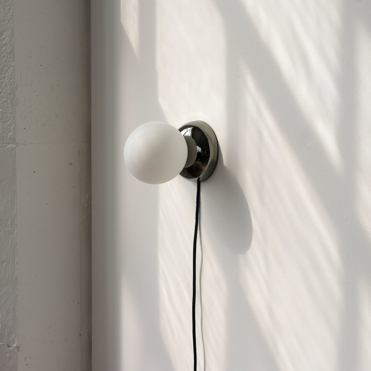 Wall-mounted sconce with a round black metal base and a frosted white glass globe, casting a soft shadow on a sunlit white wall.